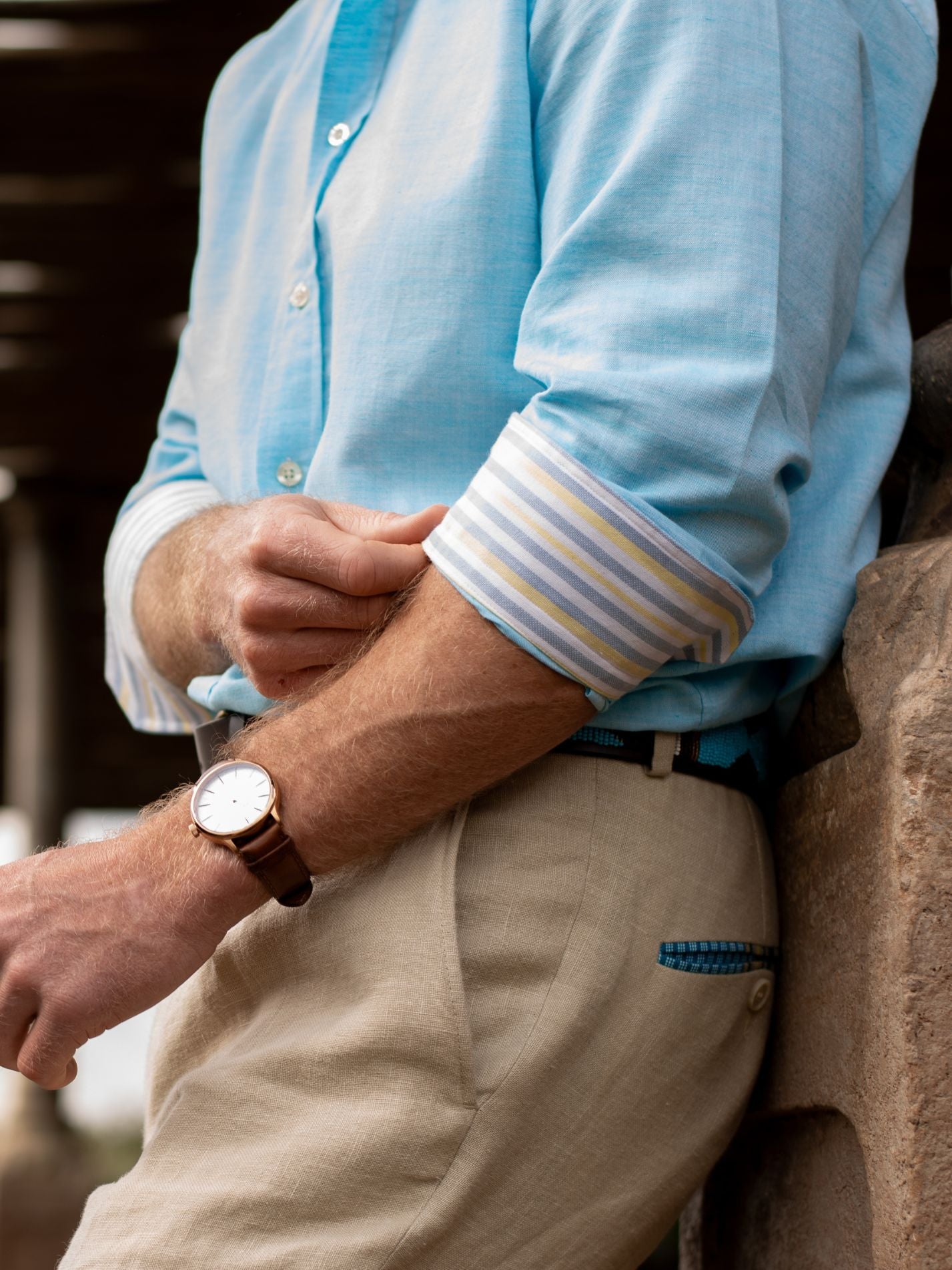 Person wearing a light blue shirt with striped cuffs and beige pants, sitting on a stone wall.