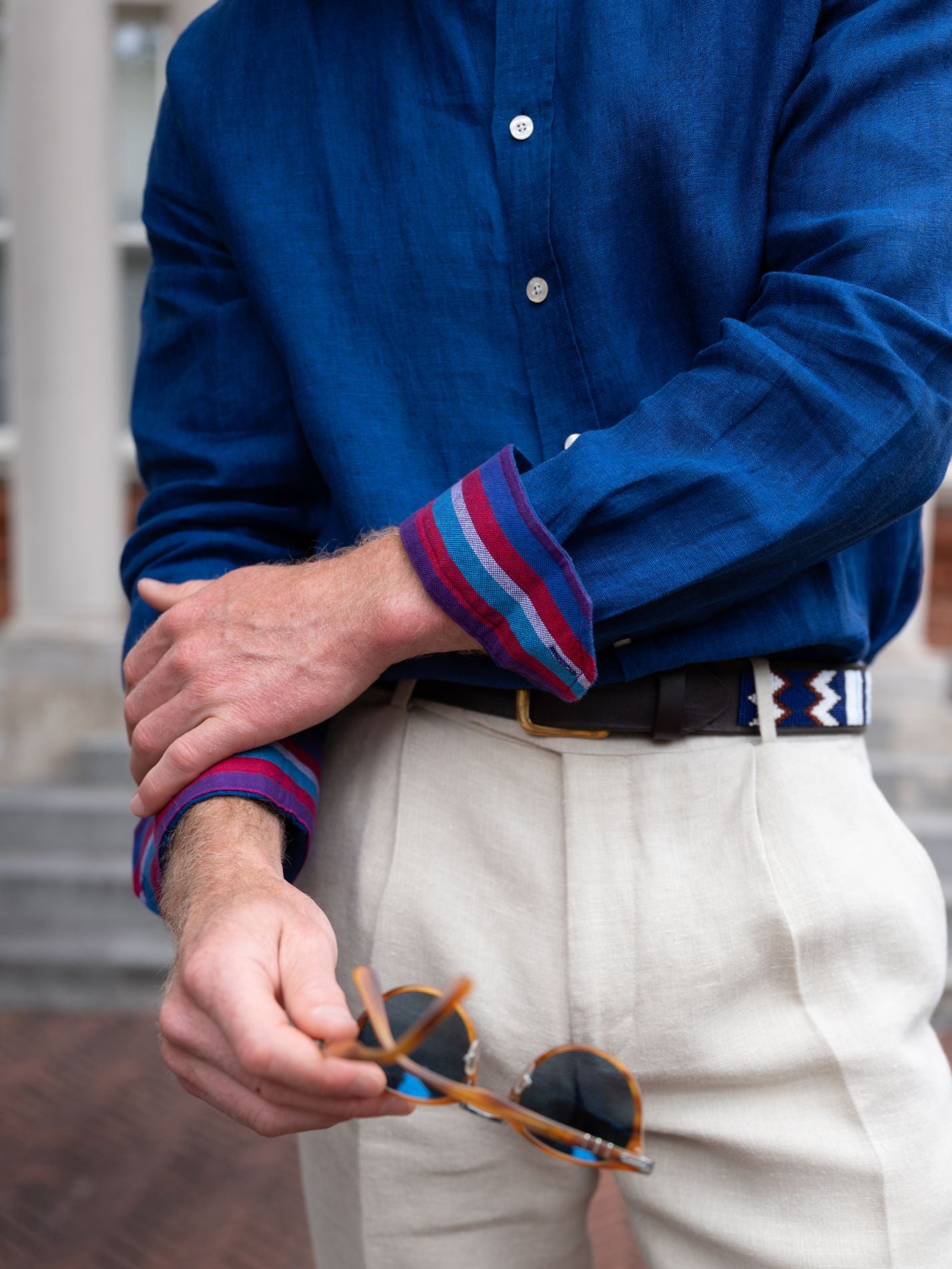 Person wearing a blue shirt with rolled-up sleeves and white pants, holding sunglasses.