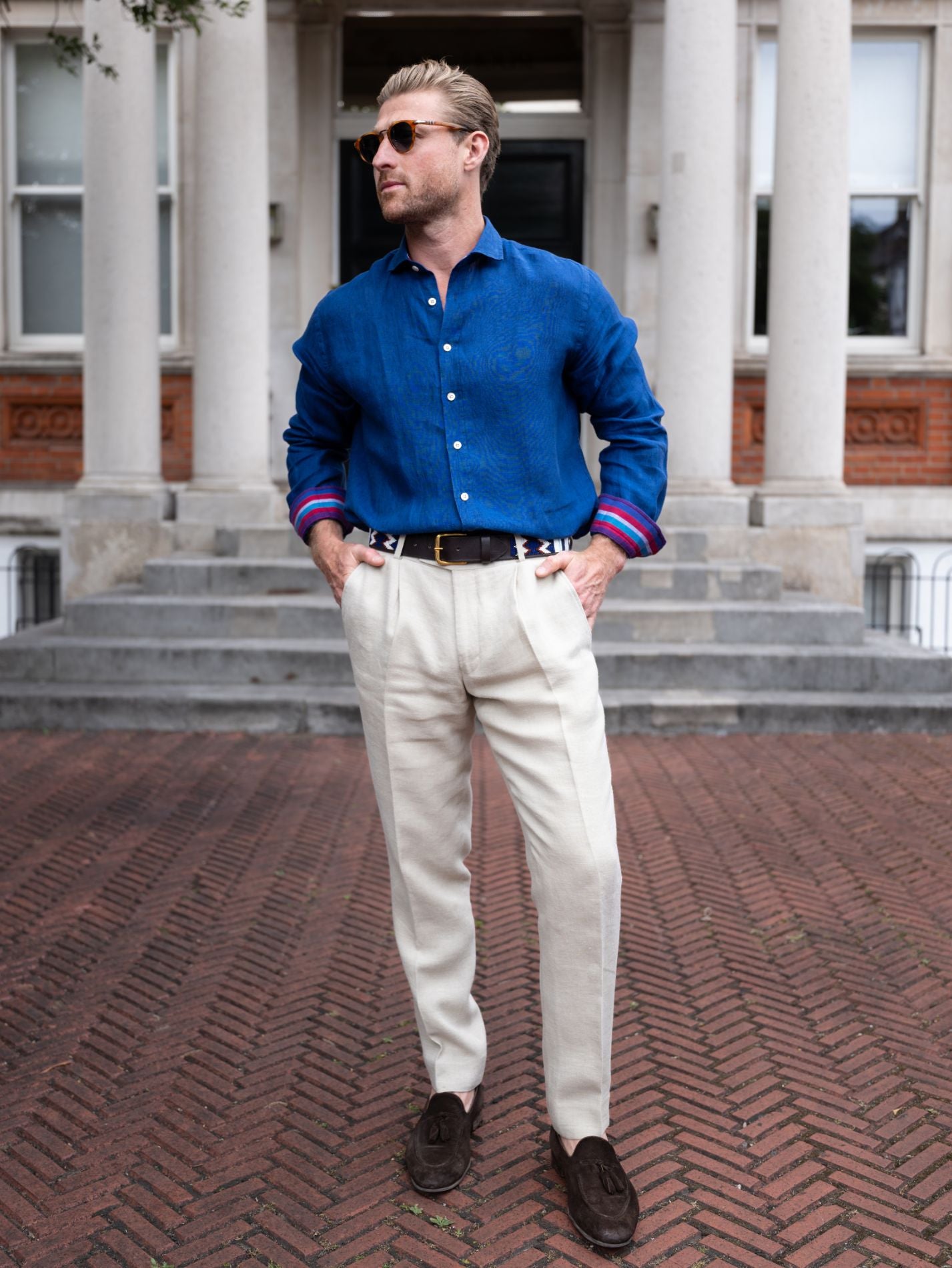 Man wearing a blue shirt with colorful trim, white pants, and dark shoes standing on a brick pavement.
