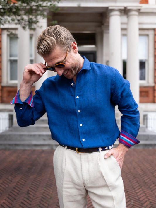 Man wearing a blue shirt with rolled-up sleeves and white pants, standing in front of a building.